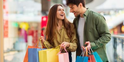 Joyful Couple On Shopping Laughing Holding Colorful Shopper Bags Buying New Clothes In Modern Hypermarket On Weekend. Happy Customers Spending Weekend In Mall. Great Sales Concept